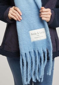 Écharpe en laine bleu clair avec des bords frangés et une étiquette blanche affichant "Andy & Lucy Paris Wrapped in Love". Texture douce et design léger.