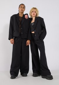 Zwarte oversized blazers met knopen aan de voorkant, gecombineerd met brede broekspijpen. De outfits hebben een zachte textuur en een hedendaagse silhouet.
