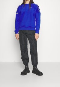Personne portant un sweat à capuche bleu avec des logos sur les manches et un pantalon de jogging noir avec des bottes épaisses noires.