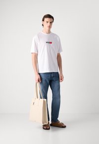 Bala balta kokvilnas T-kreklā ar uzrakstu "HUG" un sirds grafiku, zili denimu džinsi, beiga krāsas somiņa un brūni slidenskapī. Vienkārša, ikdienišķa apģērba kombinācija.