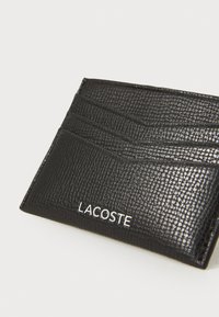 Sort tekstureret læderkortholder med multiple kortpladser og et hvidt "LACOSTE" logo på forsiden. Rektangulær form.