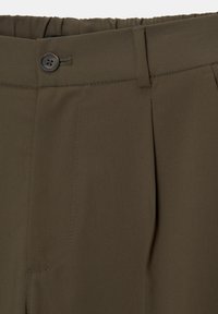 Gros plan sur la ceinture d’un pantalon vert olive avec fermeture à bouton, passant de ceinture et détail de tissu plissé.
