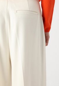 Wide-leg trousers in cream fabric with a smooth texture, featuring a single back pocket and minimal stitching detail, paired with an orange top.
