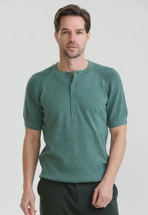 Mann mit kurzen braunen Haaren, der ein grünes kurzärmeliges Henley-Shirt und dunkle Hose trägt, steht vor einem schlichten hellen Hintergrund.