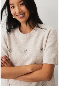 Kurzärmeliger, hellbeiger Wollpullover mit weicher Textur. Verziert mit floralen Perlenapplikationen in Silber und einem Rundhalsausschnitt.