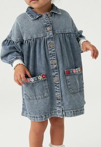 Jeanskleid mit langen Ärmeln, Knopfleiste und ausgestellter Form. Verfügt über dekorative florale Applikationen auf den Taschen. Hellblaue Farbe und weiche Textur.