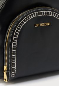 Μαύρη δερμάτινη τσάντα με στρογγυλεμένη θήκη με φερμουάρ, διακοσμημένη με χρυσές λεπτομέρειες και λευκά ραφικά. Εμφανίζεται το όνομα της μάρκας "LOVE MOSCHINO".