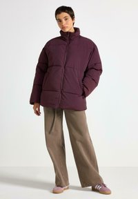 Giubbotto in piumino color bordeaux con colletto alto e cuciture visibili. Presenta due tasche frontali, abbinato a pantaloni larghi color khaki e sneakers rosa.