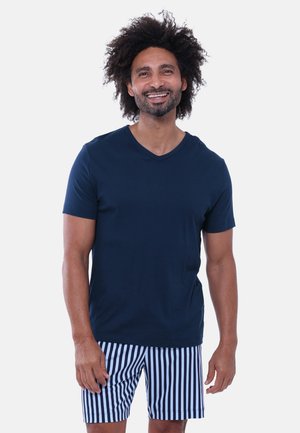 Uomo sorridente con capelli ricci che indossa una camicia blu navy a maniche corte e pantaloncini a righe verticali blu e bianche, in piedi davanti a uno sfondo semplice.