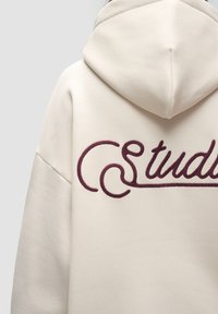 Beżowa bluza z kapturem z dużym, ciemno-burgundowym haftowanym napisem "Studio" na plecach, o miękkiej teksturze i z kapturem regulowanym sznurkiem.