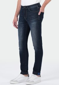 Homme portant un jean slim bleu foncé avec les poignets roulés et des baskets blanches, se tenant avec une main dans la poche contre un fond blanc.