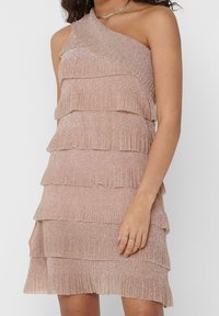 Kvinna iklädd en ärmlös, enaxlad, tärnad, glittrande beige klänning med plisserade lager och silverringar på sin högra hand.