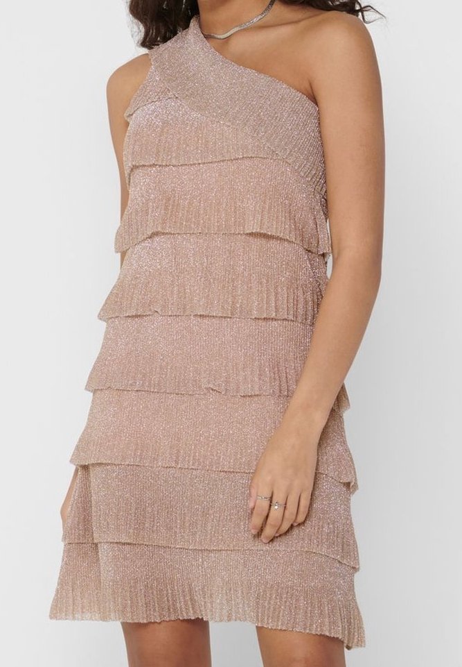 Kvinna iklädd en ärmlös, enaxlad, tärnad, glittrande beige klänning med plisserade lager och silverringar på sin högra hand.