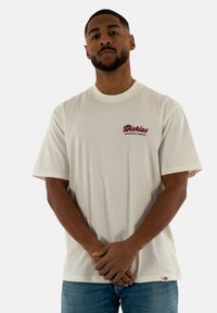 Kremo spalvos trumpomis rankovėmis marškinėliai su raudonu "Dickies" logotipu. Minkšta medvilnė, laisvas kirpimas, apvali iškirptė ir šoninis etiketės detalė.