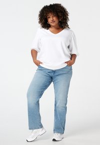 Femme aux cheveux bouclés se tenant avec les mains dans les poches, portant une blouse blanche, un jean bleu clair et des baskets blanches devant un fond blanc.