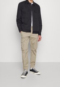 Man in een zwart knoopjasje, witte shirt, beige cargobroek met zakken, en zwart-witte sneakers, staande voor een effen achtergrond.
