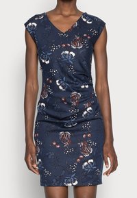 Ärmlös marinblå klänning med V-ringning och blommönster i vitt, blått och brunt, bärs av en person med mörk hud.