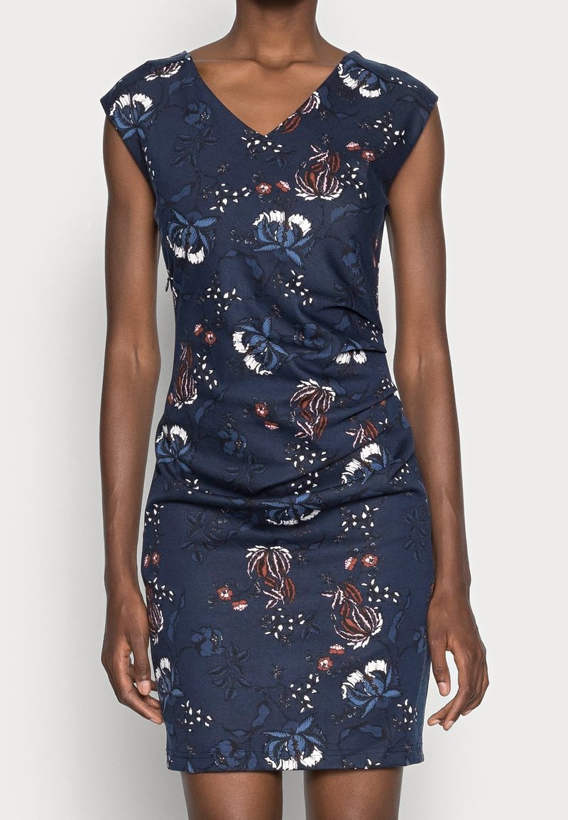 Ärmlös marinblå klänning med V-ringning och blommönster i vitt, blått och brunt, bärs av en person med mörk hud.