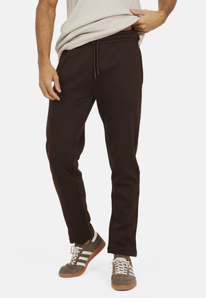 Man draagt donkerbruine joggerbroek met trekkoord en grijze sneakers met witte strepen, tilt beige shirt lichtelijk op.