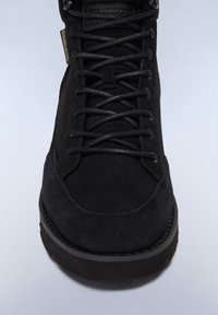 Botín de ante negro con parte superior texturizada, cordones redondos y suela de goma en contraste. Diseño minimalista sin marcas visibles.