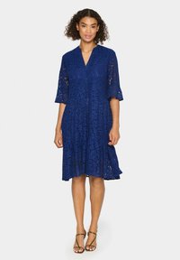 Rochie din dantelă bleu marin cu decolteu în V și mâneci trei sferturi. Poate fi purtată cu un tiv evazat și un model floral pe întreaga suprafață. Combinată cu sandale cu bretele.