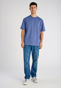 T-shirt in cotone blu con maniche corte, scollo rotondo, indossato con jeans di denim blu e sneakers bianche con accenti di colore. Sfondo da studio.