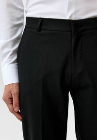 Zwarte, op maat gemaakte broeken van een gladde stof met een slim fit. Voorzien van een platte voorkant, riemlussen en zijzakken, gestyled met een wit shirt.