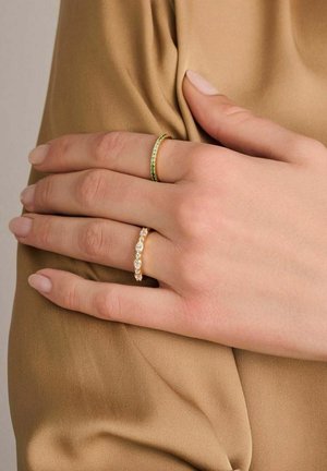 To ringe på en hånd: den ene er en slank guld med grønne ædelsten, og den anden er en delikat guld ring med klare, ovale ædelsten.