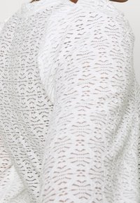 Närbild av ett vitt strukturerat stickat tyg med ett geometriskt, utskuret mönster på en ärm och axel.