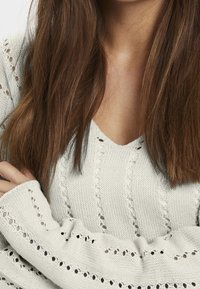 Light gray knitted sweater with a V-neck, featuring openwork patterns and ribbed cuffs. Textured fabric allows for breathability.