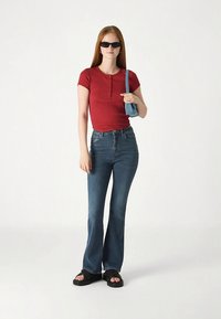 Κόκκινη μπλούζα με κοντά μανίκια και κουμπιά, συνδυασμένη με καμπανέ μπλε τζιν και μαύρα σανδάλια. Αξεσουάρ: μικρή μπλε τσάντα και γυαλιά ηλίου.