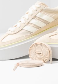 Béžové atletické boty s nubukovým svrškem a síťovinovými detaily, s třemi bílými pruhy. Obsahují stočené béžové tkaničky vedle boty.