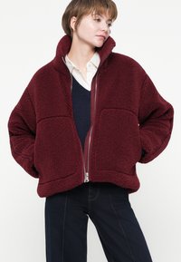 Femeie cu părul scurt, purtând o jachetă oversized din sherpa de culoare burgundă peste o cămașă albă și un pulover bleu, cu mâinile în buzunare, privind spre lateral.