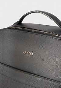 Crna kožna torba s teksturom, s gornjom držačem, vidljivim zatvaračem i zlatnim logotipom "Lancel Paris" na prednjoj površini.