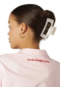 Camicia rosa con testo ricamato "Non la mamma media" in rosso. Capelli acconciati in uno chignon con una grande molletta rettangolare color crema.