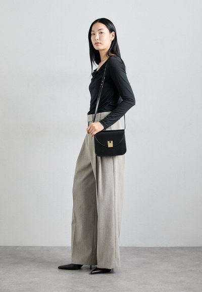 Black leather crossbody bag with gold hardware, sleek design, and curved flap. Model wears fitted long-sleeve top and light grey wide-leg trousers.