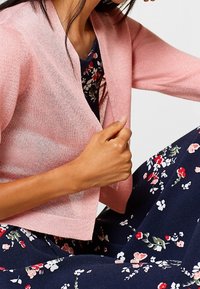 Krátký růžový pletený kardigan s měkkou texturou, s třičtvrtečními rukávy, nošený přes tmavé šaty s květinovým vzorem.