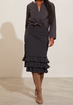 Frau trägt ein dunkles, langärmliges, gebundenes Hemd, einen schwarzen, gerafften Midirock und hellbraune Wildlederstiefel bis zum Knie, steht in einem Innenraum auf einem hellen Boden.