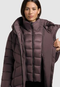 Pufasta jakna v temno mauvni barvi, s prešito zasnovo, visokim ovratnikom in dvema zadrgama. Material se zdi mehak in izoliran.