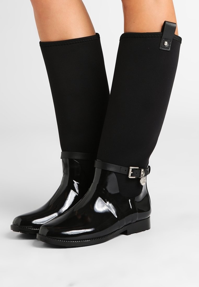 Bottes noires jusqu'aux genoux avec section inférieure brillante et partie supérieure mate, dotées d'une sangle avec boucle et pendentif autour de la cheville, portées sur des jambes nues.