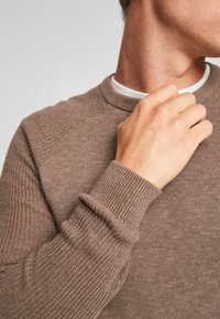 Brauner Strickpullover mit gerippten Bündchen und Ausschnitt, der ein strukturiertes Muster an den Schultern aufweist. Insgesamt glatter Stoff, lässiges Design.