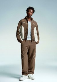 Brown jacket with light grey accents and white stitching, paired with matching brown pants and white sneakers, all in a relaxed fit.