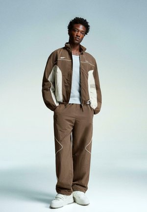 Bruine jas met lichtgrijze accenten en witte naden, gecombineerd met bijpassende bruine broek en witte sneakers, allemaal in een ontspannen pasvorm.