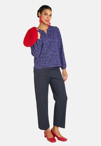 Frau in bluse mit blauem Muster und langen Ärmeln, dunkler Hose, roten herzförmigen Ohrringen, die eine rote Baskenmütze hält, und roten Absätzen vor weißem Hintergrund.