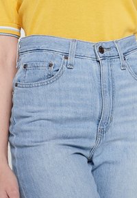 Jeans de tiro alto en azul claro con un diseño de cinco bolsillos, sutil deslavado y costuras contrastantes. Combinados con una camisa amarilla.