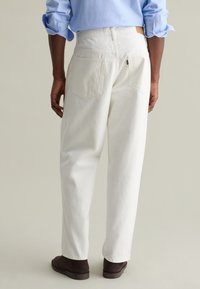 Pantalones de algodón blancos con un corte relajado, que cuentan con dos bolsillos traseros y un detalle de cuero marrón en la cintura. Calzado de ante marrón visible.
