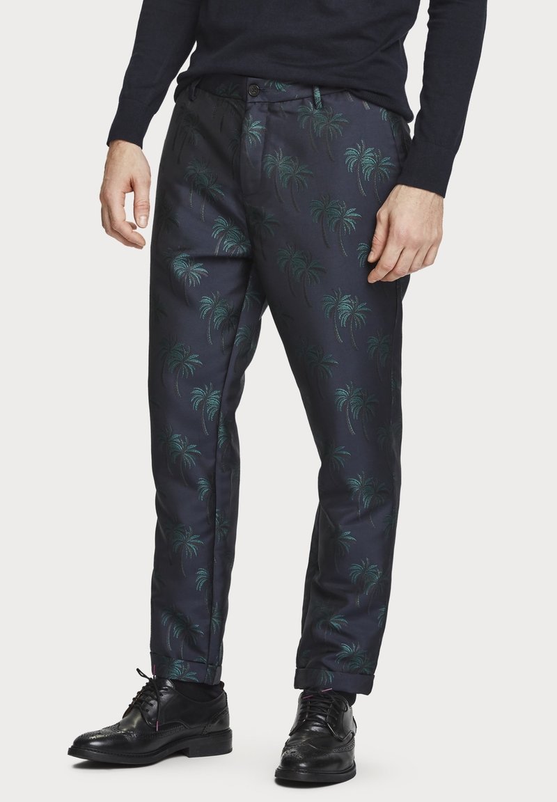 Mann in dunklen Hosen mit grünen Palmenmustern, schwarzen Schuhen und einem langen, dunklen Oberteil, stehend vor einem weißen Hintergrund.