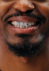 Primo piano di un sorriso con grillz dentali. I grillz mostrano vari strass blu e forme geometriche su denti bianchi, in contrasto con una barba scura.