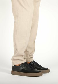 Sneakers in pelle nera con dettagli in suede, suola in gomma color nocciola e lacci neri. Indossati con pantaloni beige chiaro su misura.