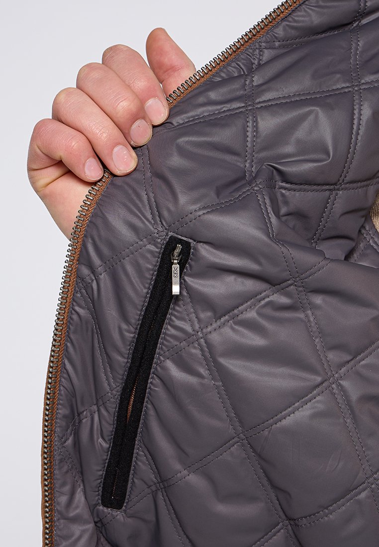 Wnętrze pikowanej szarej kurtki z widoczną kieszenią na zamek błyskawiczny, wykończoną czarną tkaniną. Brązowe wykończenie na zamku podkreśla funkcjonalność.